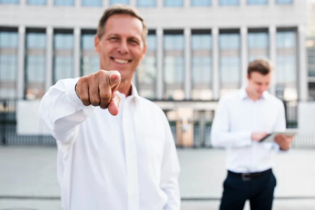 Hombre señalando al frente con una sonrisa, al fondo un condominio con una persona con dispositivo móvil.