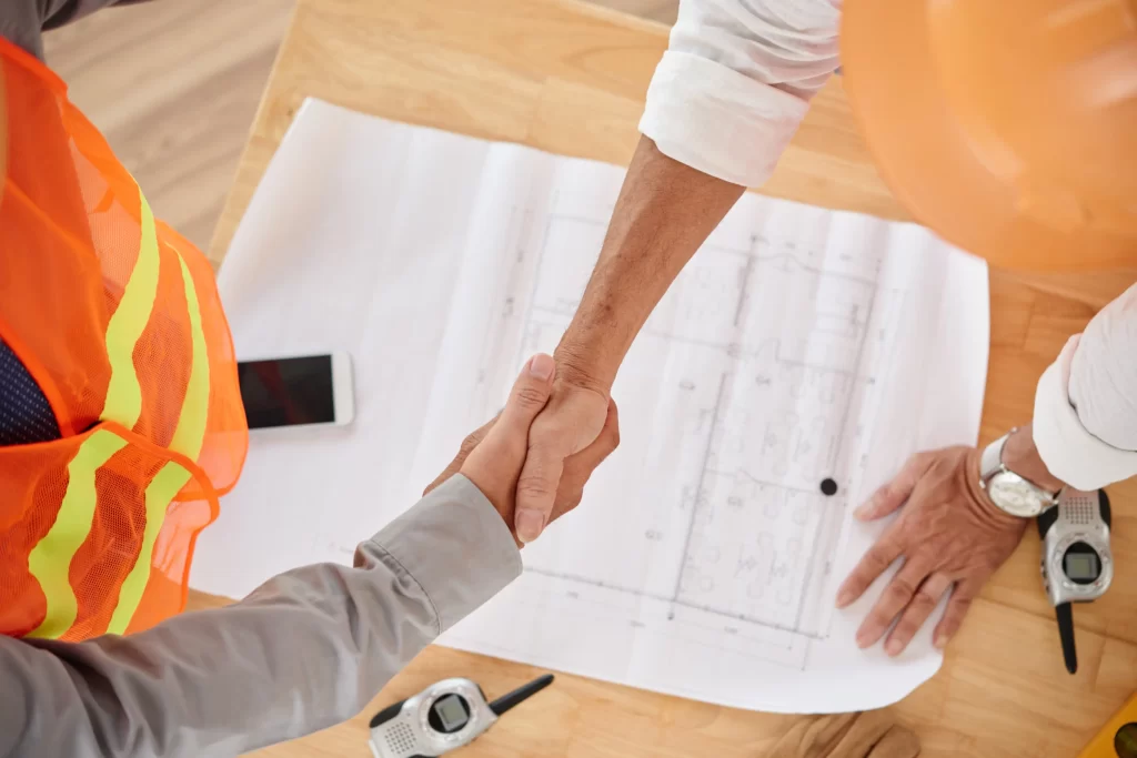 Firme apretón de manos entre ingenieros con papeles de construcción de fondo.